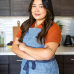 Dara Yu standing confidently in a kitchen, wearing a denim apron over an orange shirt, with arms crossed and a slight smile.