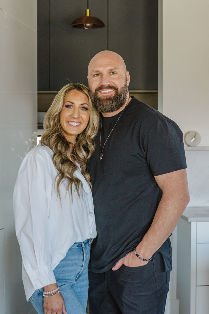 Janae and Kevin Cox smiling together in a modern kitchen, Janae wearing a white blouse and jeans, Kevin in a black t-shirt with his arm around her.