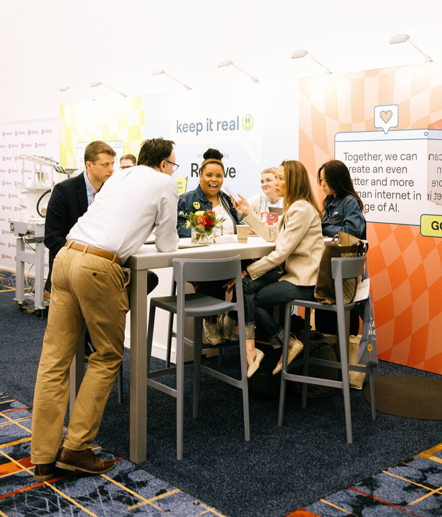 A group of people sit and stand around a tall table having a lively conversation at a conference booth with colorful signage reading “keep it real” and “Together, we can create an even better and more human internet in the age of AI.”