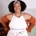 A woman in an apron stands smiling in front of an oven with red knobs.