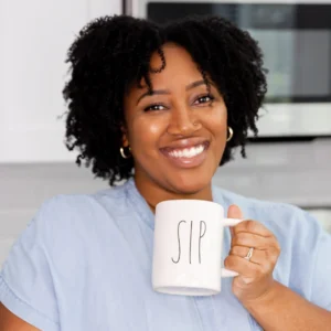 Jessica Hylton smiling and holding a white mug that says “SIP” while standing in a bright kitchen.