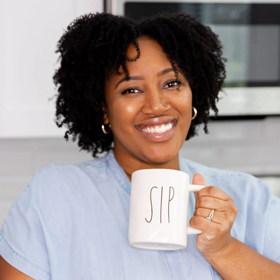 Jessica Hylton smiling and holding a white mug that says “SIP” while standing in a bright kitchen.