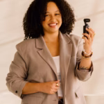 Natasha Pierre smiles while holding a small handheld camera, wearing a tan blazer over a white top against a softly lit neutral background.