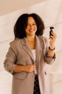 Natasha Pierre smiles while holding a small handheld camera, wearing a tan blazer over a white top against a softly lit neutral background.
