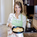 Laura Ashley Johnson smiling in a kitchen while holding a cast-iron skillet of freshly baked biscuits, wearing a light green Williams Sonoma apron over a white blouse.