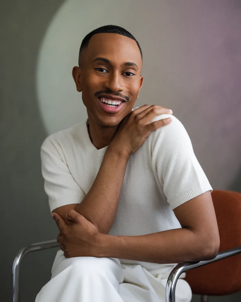 Kalen Allen smiling and seated in a rust-colored chair, wearing a cream short-sleeve knit top and white pants against a soft neutral background.