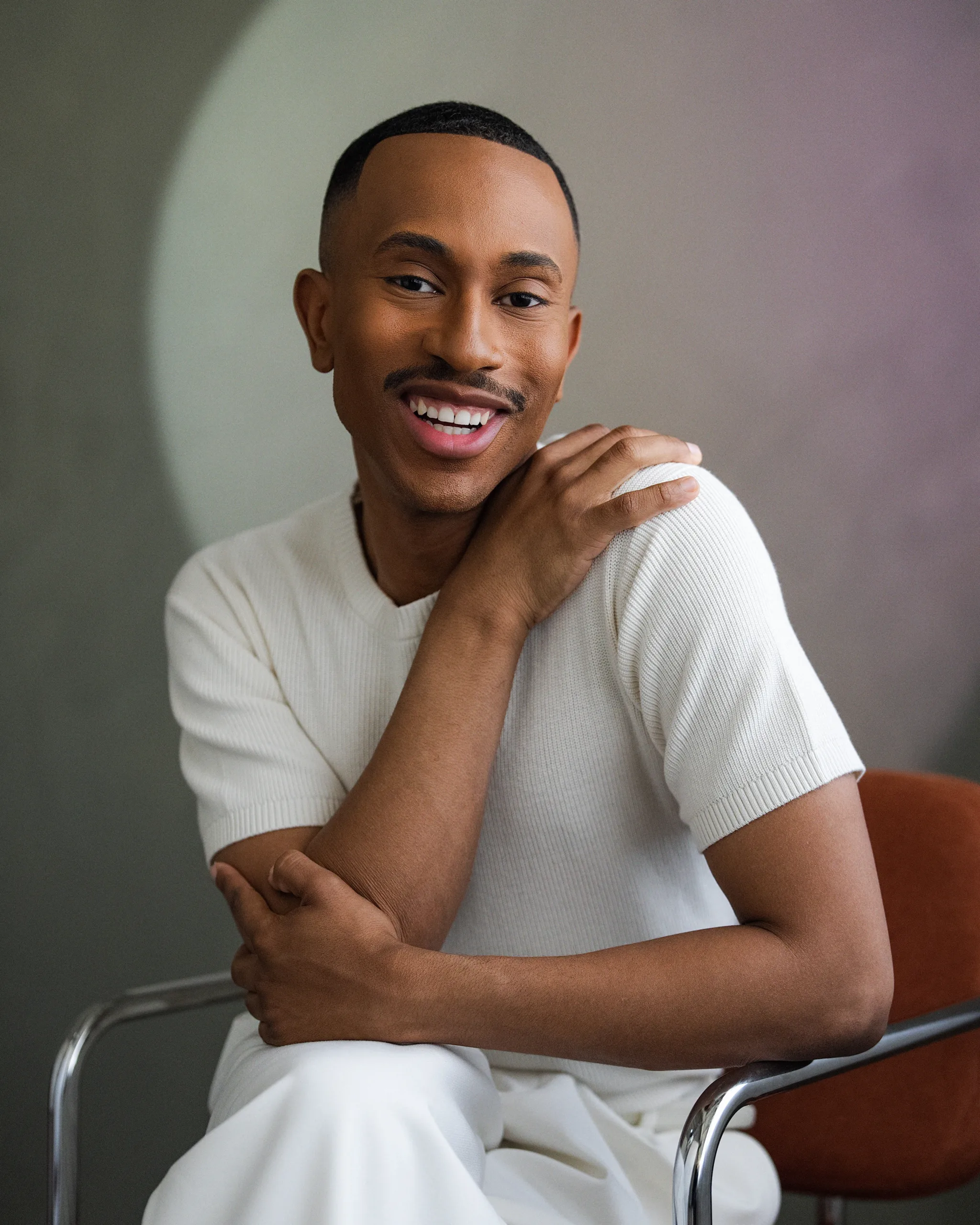 Kalen Allen smiling and seated in a rust-colored chair, wearing a cream short-sleeve knit top and white pants against a soft neutral background.