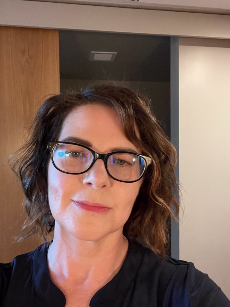 Headshot of Heather Wilkins, a woman with short wavy brown hair and black glasses, wearing a black top.