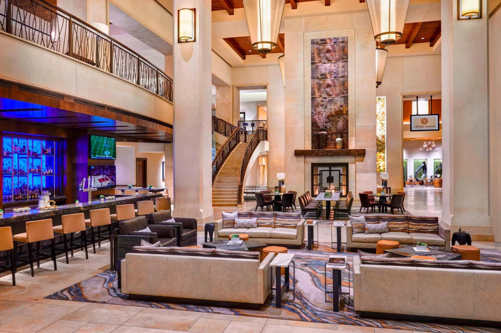 Spacious lobby of the JW Marriott San Antonio Hill Country Resort & Spa featuring high ceilings, a central fireplace, modern lounge seating, a bar with blue backlighting, and a sweeping staircase framed by warm stone and wood architectural details.