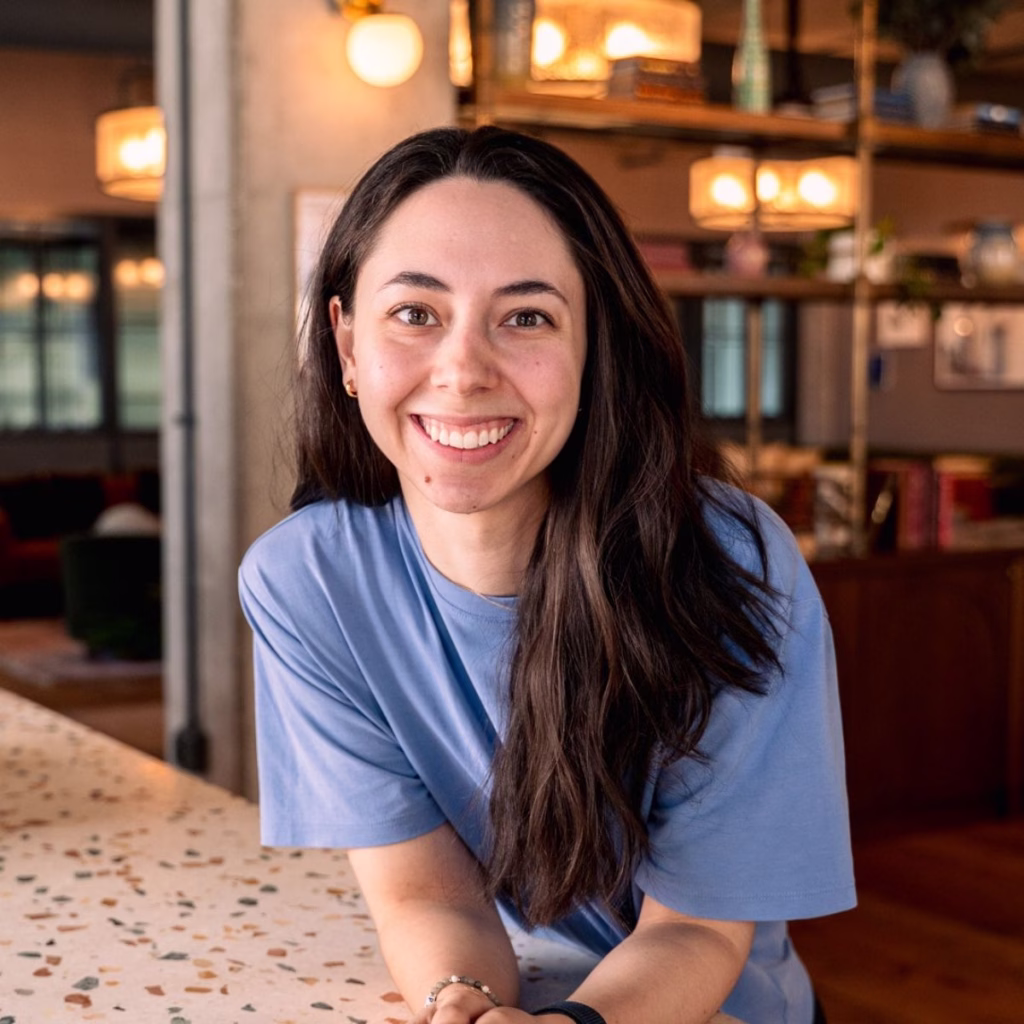 A woman in a blue t-shirt with long brown hair stands smiling in her kitchen. She leans on a kitchen island and the lighting is warm and calm.