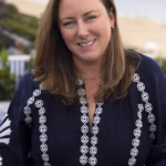 Portrait of Amanda Martin smiling outdoors near the coast, wearing a navy blue embroidered blouse, with the ocean and sandy beach softly blurred in the background.