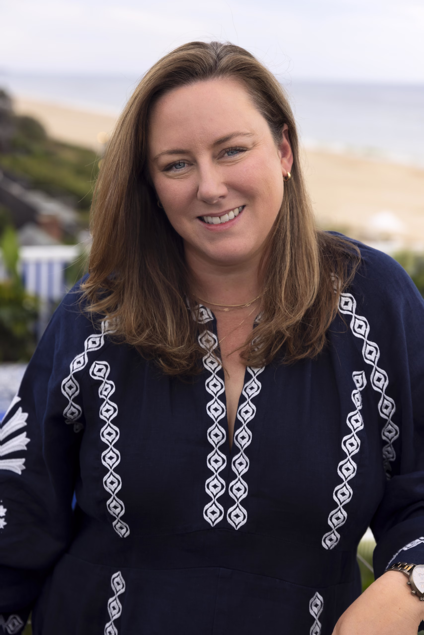 Portrait of Amanda Martin smiling outdoors near the coast, wearing a navy blue embroidered blouse, with the ocean and sandy beach softly blurred in the background.