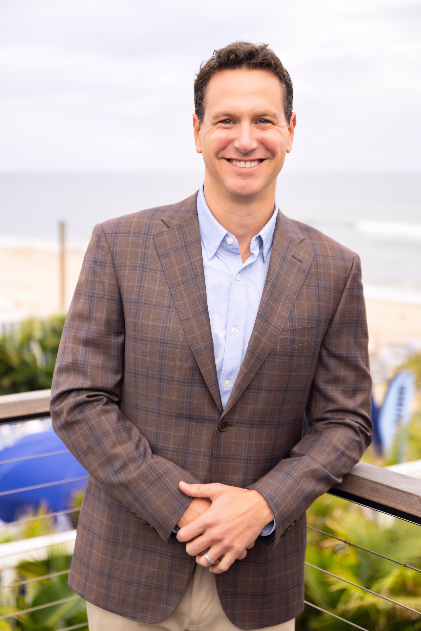 Eric Hochberger smiles at the camera while standing outdoors near the ocean, wearing a brown plaid blazer over a light blue button-down shirt, with his hands resting on a railing and a beachside background softly blurred behind him.
