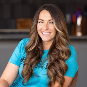 Headshot of Susie Bulloch smiling indoors, wearing a teal T-shirt, with long wavy brown hair and a softly blurred background.