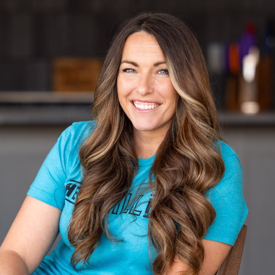 Headshot of Susie Bulloch smiling indoors, wearing a teal T-shirt, with long wavy brown hair and a softly blurred background.