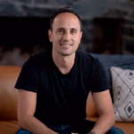 Ben Jabbawy seated on a brown leather couch, smiling at the camera, wearing a black T-shirt and jeans. He is sitting with hands clasped, with a stone fireplace and patterned pillow in the background.