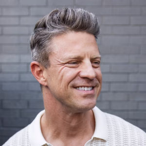Matt Molen smiling and looking to the side, with short salt-and-pepper hair styled up, wearing a light-colored textured polo shirt, photographed in front of a gray brick wall.