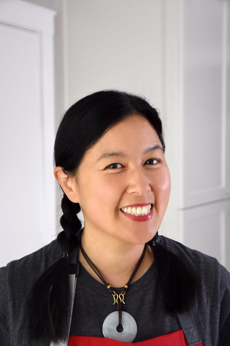 Michelle Tam smiling at the camera, wearing a dark shirt and apron, with long black hair in braided pigtails and a round pendant necklace, photographed against a light neutral background.