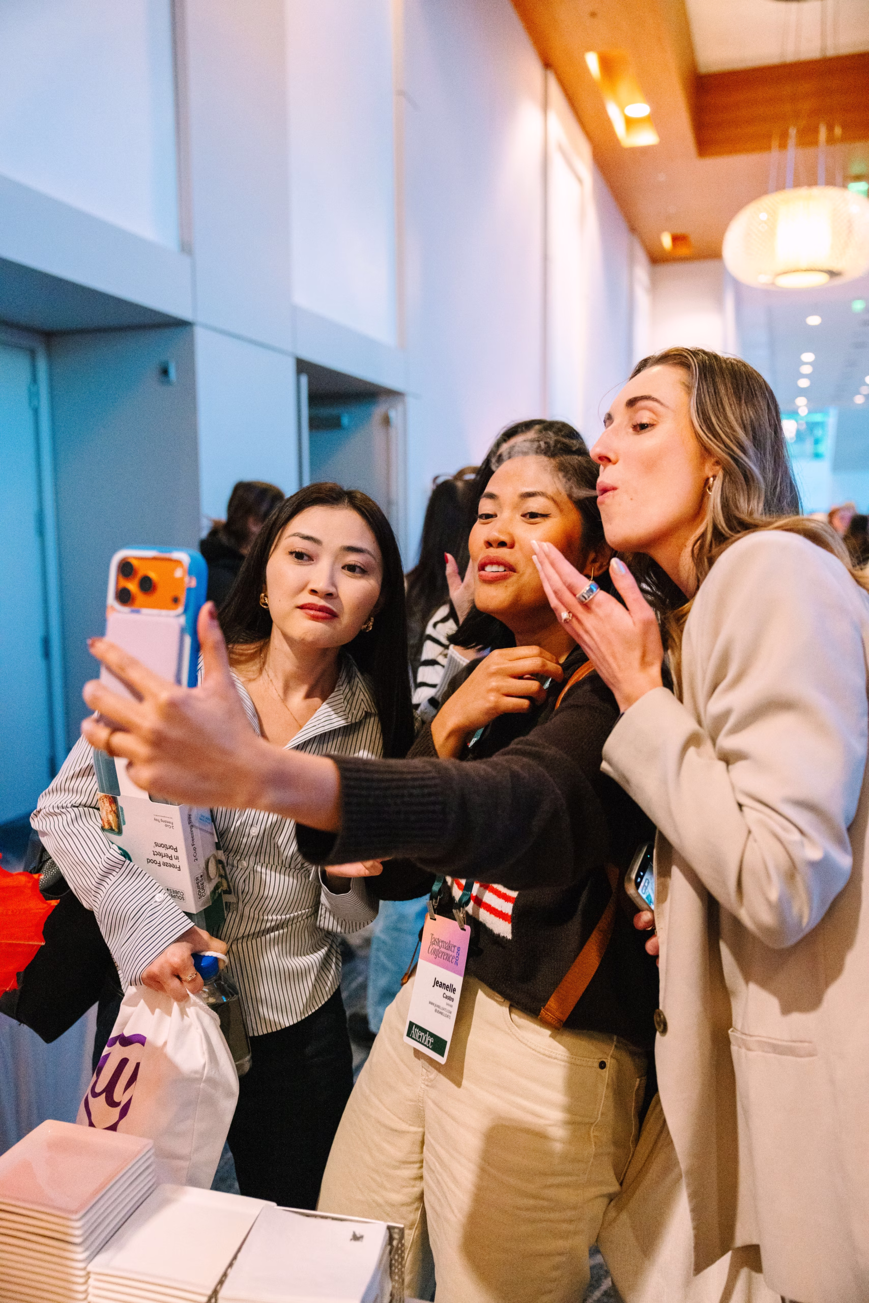 Three women wearing conference badges take a selfie together in a hallway, smiling and posing for the camera during an event.