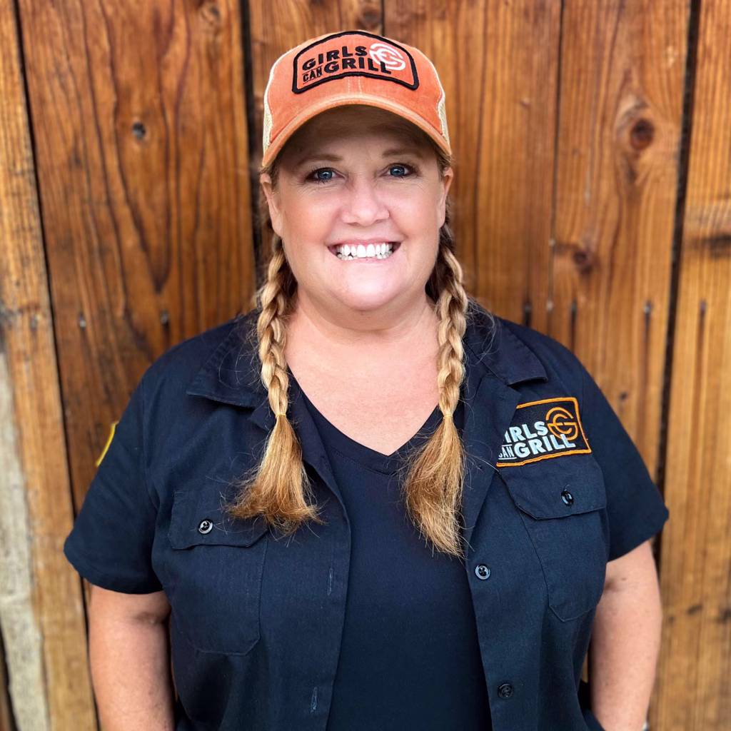 Smiling Christie Vanover with blonde hair in two braids, wearing a Girls Can Grill baseball cap and black branded shirt, standing in front of a wooden wall.