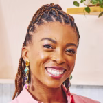 Jenné Claiborne smiling at the camera with braided hair pulled back, wearing colorful gemstone drop earrings and a pink blouse, photographed in a bright kitchen setting.