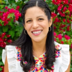 Headshot of Dora Ramirez smiling outdoors in front of vibrant pink flowers, wearing a white blouse with colorful embroidered detailing and silver hoop earrings.
