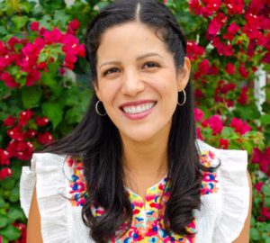 Headshot of Dora Ramirez smiling outdoors in front of vibrant pink flowers, wearing a white blouse with colorful embroidered detailing and silver hoop earrings.