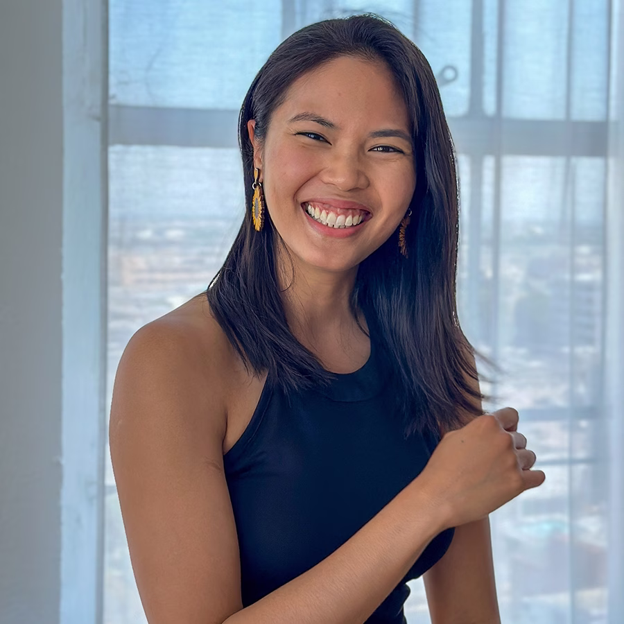 Jeanelle Castro smiling, wearing a sleeveless black top and gold earrings, standing indoors in front of a large window with a softly blurred city view.