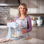 Sally McKenney smiling while using a light blue stand mixer in a bright home kitchen, wearing a floral apron and preparing ingredients on a marble countertop.