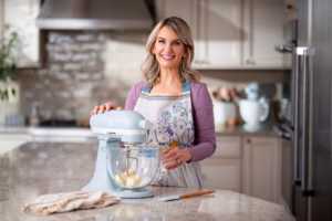 Sally McKenney smiling while using a light blue stand mixer in a bright home kitchen, wearing a floral apron and preparing ingredients on a marble countertop.