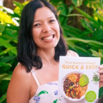 Toni Okamoto smiling outdoors in front of lush green plants, holding her cookbook “Plant-Based on a Budget: Quick & Easy,” which features a bowl of colorful plant-based food on the cover.
