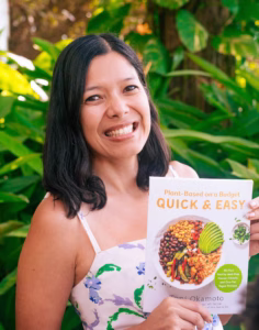 Toni Okamoto smiling outdoors in front of lush green plants, holding her cookbook “Plant-Based on a Budget: Quick & Easy,” which features a bowl of colorful plant-based food on the cover.