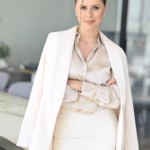 Anastasia Shtompel stands with her arms crossed in a bright office, wearing a cream satin blouse, white skirt, and white blazer draped over her shoulders.