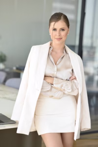 Anastasia Shtompel stands with her arms crossed in a bright office, wearing a cream satin blouse, white skirt, and white blazer draped over her shoulders.
