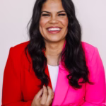 Jordan Gill smiling at the camera in a studio portrait, wearing a bold half-red, half-hot-pink blazer over a white top against a light neutral background.