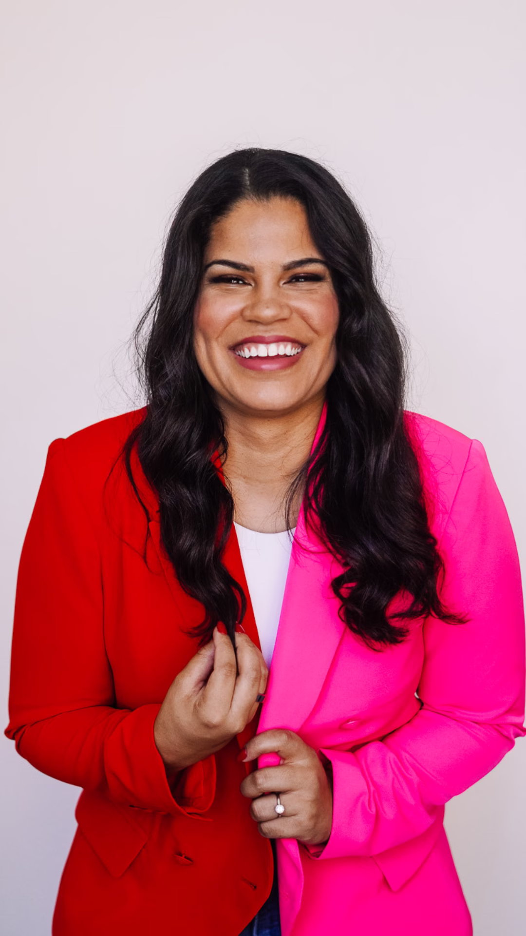 Jordan Gill smiling at the camera in a studio portrait, wearing a bold half-red, half-hot-pink blazer over a white top against a light neutral background.