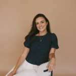 Kaitlin Janes smiles while seated in a textured chair against a warm neutral background, wearing a navy top and white pants.