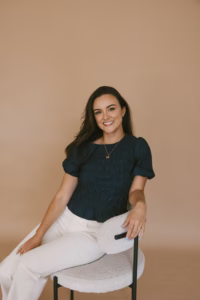Kaitlin Janes smiles while seated in a textured chair against a warm neutral background, wearing a navy top and white pants.