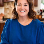 Christina Hitchcock smiles at the camera while wearing a bright blue blouse in a warmly lit kitchen.