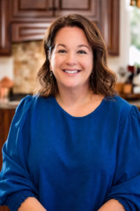 Christina Hitchcock smiles at the camera while wearing a bright blue blouse in a warmly lit kitchen.