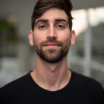 Professional headshot of Ethan Kramer wearing a black crewneck shirt, with short brown hair and a beard, smiling slightly against a softly blurred neutral background.