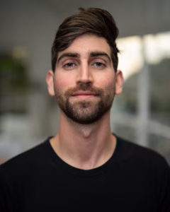 Professional headshot of Ethan Kramer wearing a black crewneck shirt, with short brown hair and a beard, smiling slightly against a softly blurred neutral background.