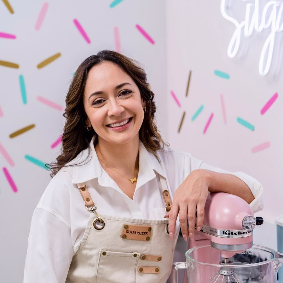 Liz Marek smiles while leaning against a pink KitchenAid mixer, wearing a white shirt and cream apron in a bright baking studio with colorful sprinkle wall decor.