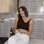 Mallory Harr smiles while sitting on a kitchen counter, holding a freshly baked cookie beside a tray of chocolate chip cookies.