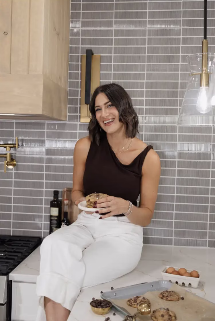 Mallory Harr smiles while sitting on a kitchen counter, holding a freshly baked cookie beside a tray of chocolate chip cookies.