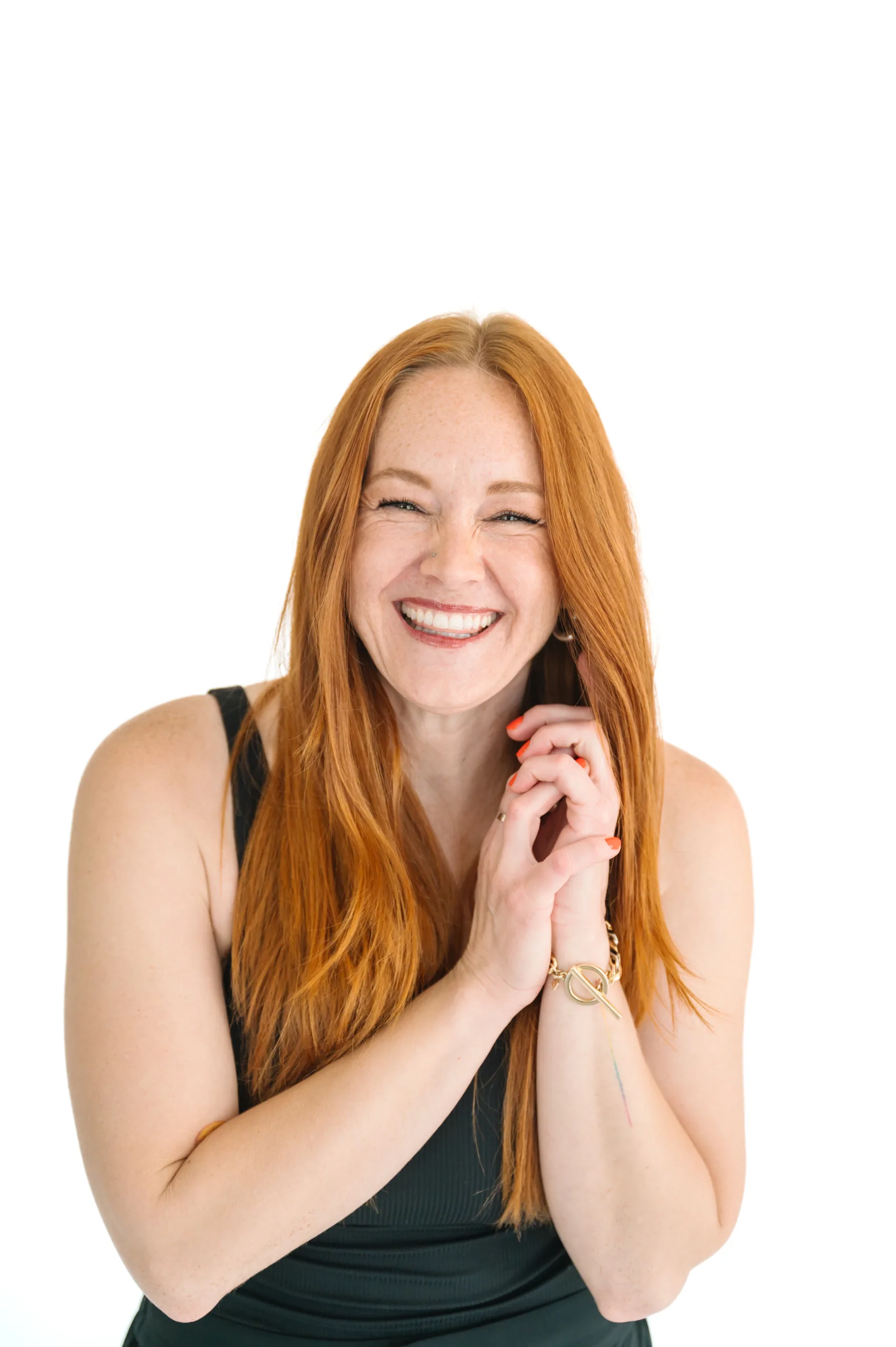 Smiling Kelli Quinn with long red hair, wearing a sleeveless black top with her hands clasped near her face, posed against a white background.