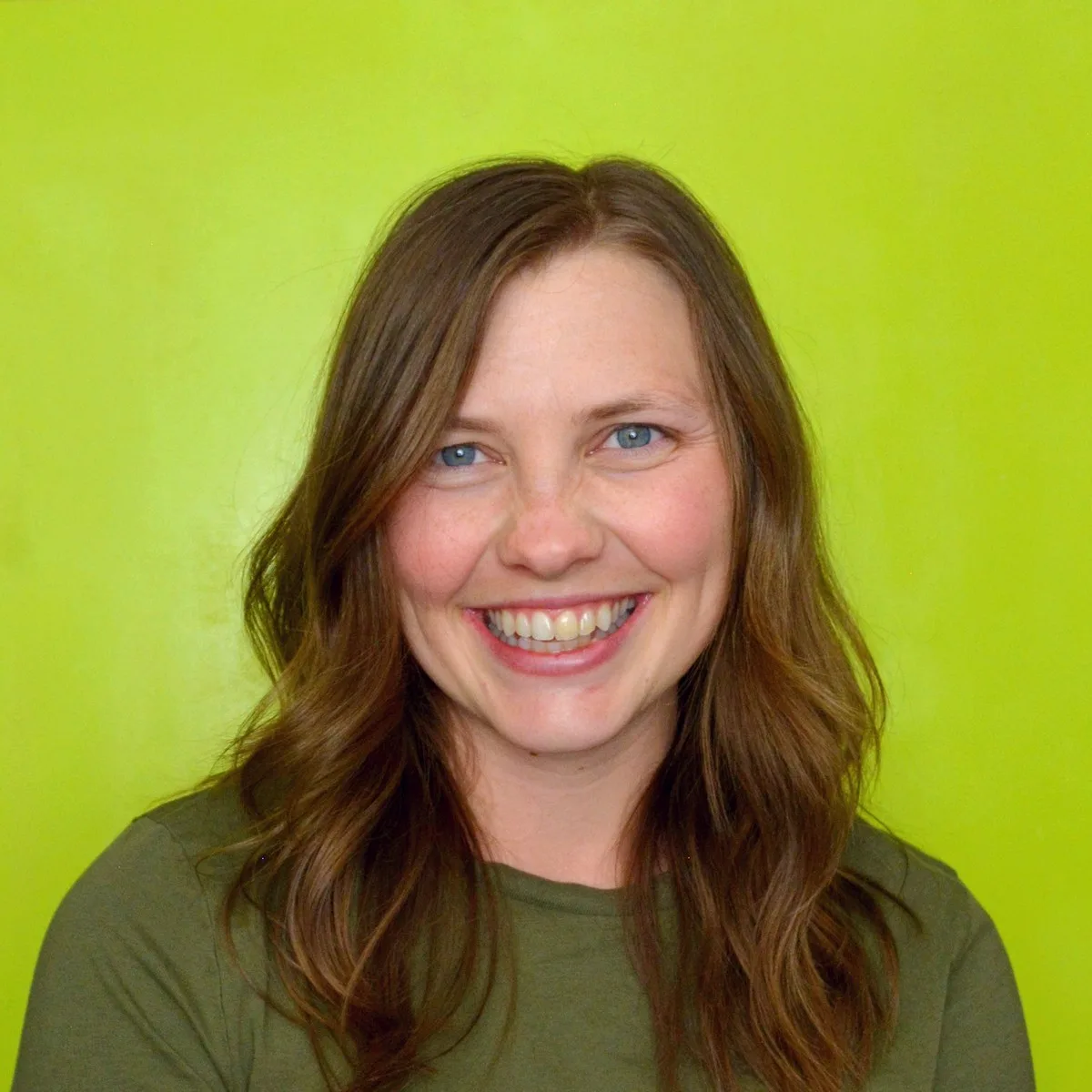 Mika Kinney smiling at the camera, with long brown hair and wearing a green top, posed against a bright green background.