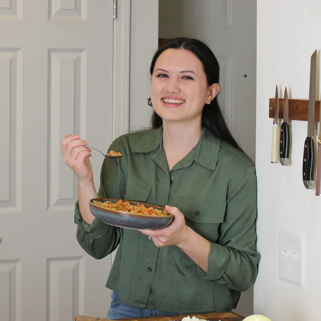 Nicole Langdon smiles at the camera while holding a bowl of food and a spoon in a bright kitchen. She wears a green button-up shirt and stands beside a wall-mounted knife rack.