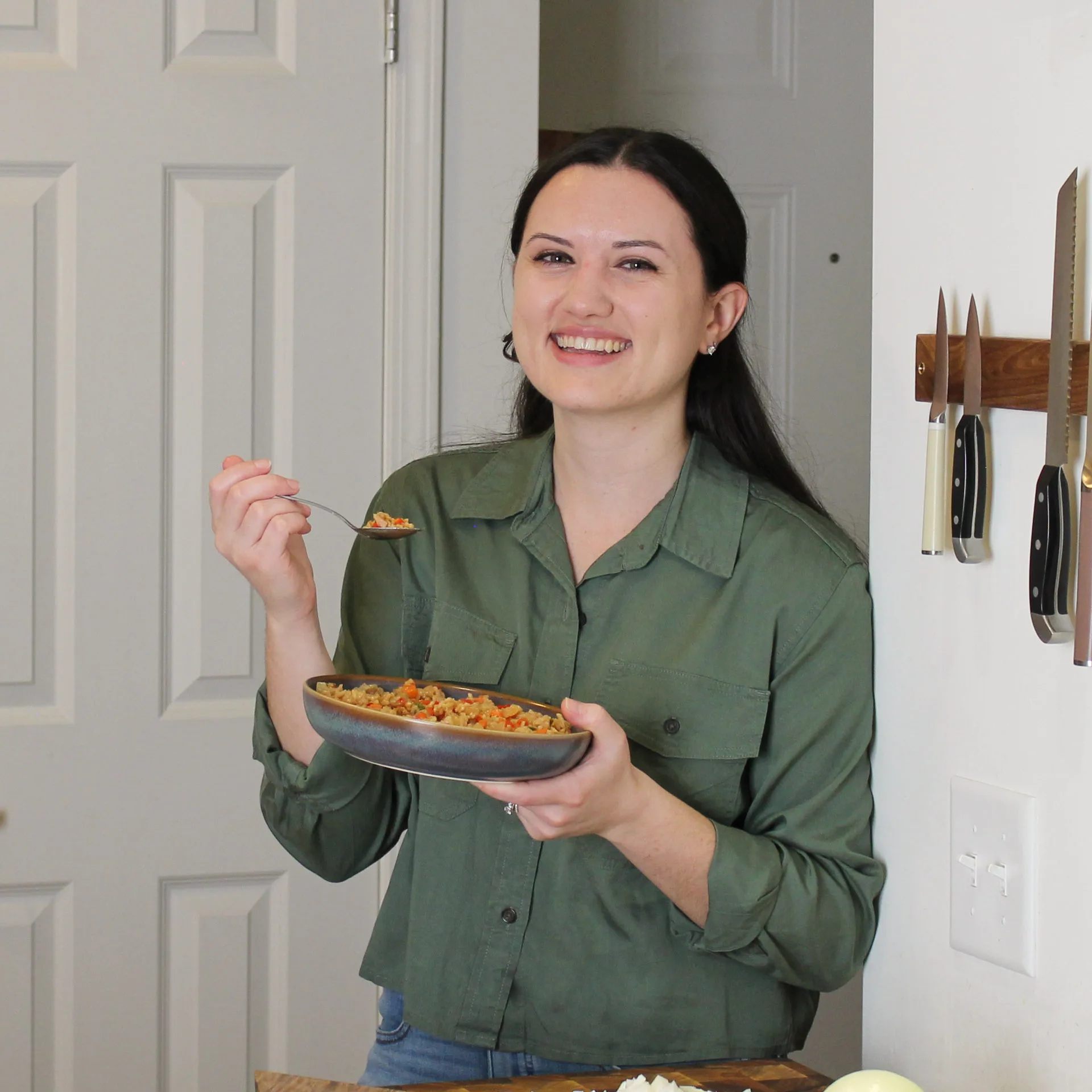 Nicole Langdon smiles at the camera while holding a bowl of food and a spoon in a bright kitchen. She wears a green button-up shirt and stands beside a wall-mounted knife rack.