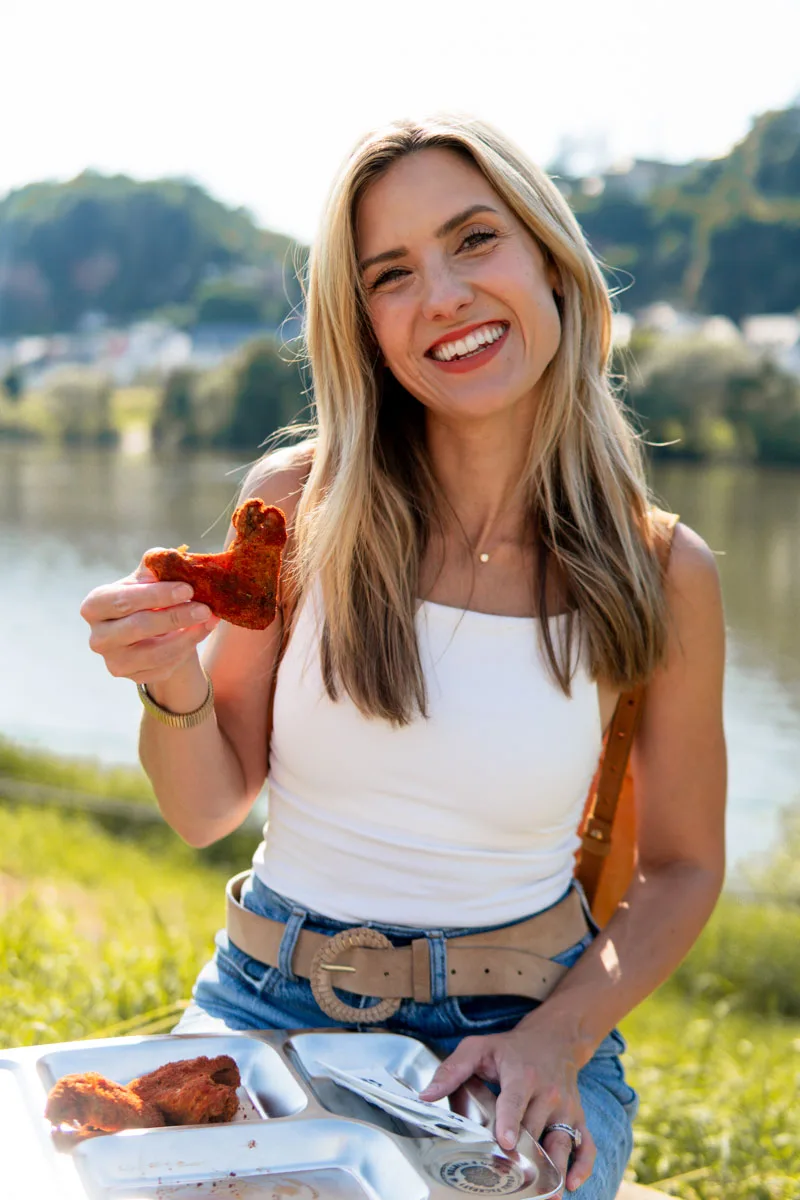 Sophie Sadler smiling outdoors by a lake while holding a piece of fried food, with a plate of food in front of her.